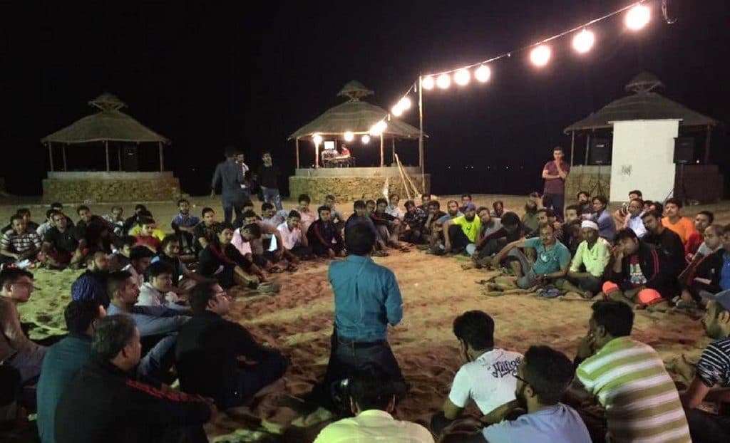 A large group sitting in a circle on sand at night, under strung lights