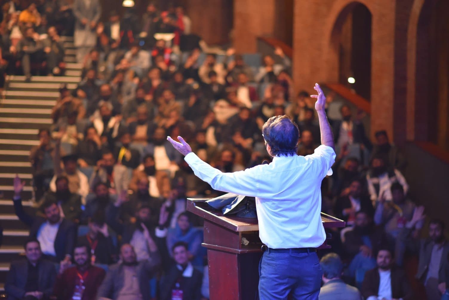 Umair Jaliawala seen from behind at the lectern, both arms raised to a packed tiered auditorium with hands going up through the rows