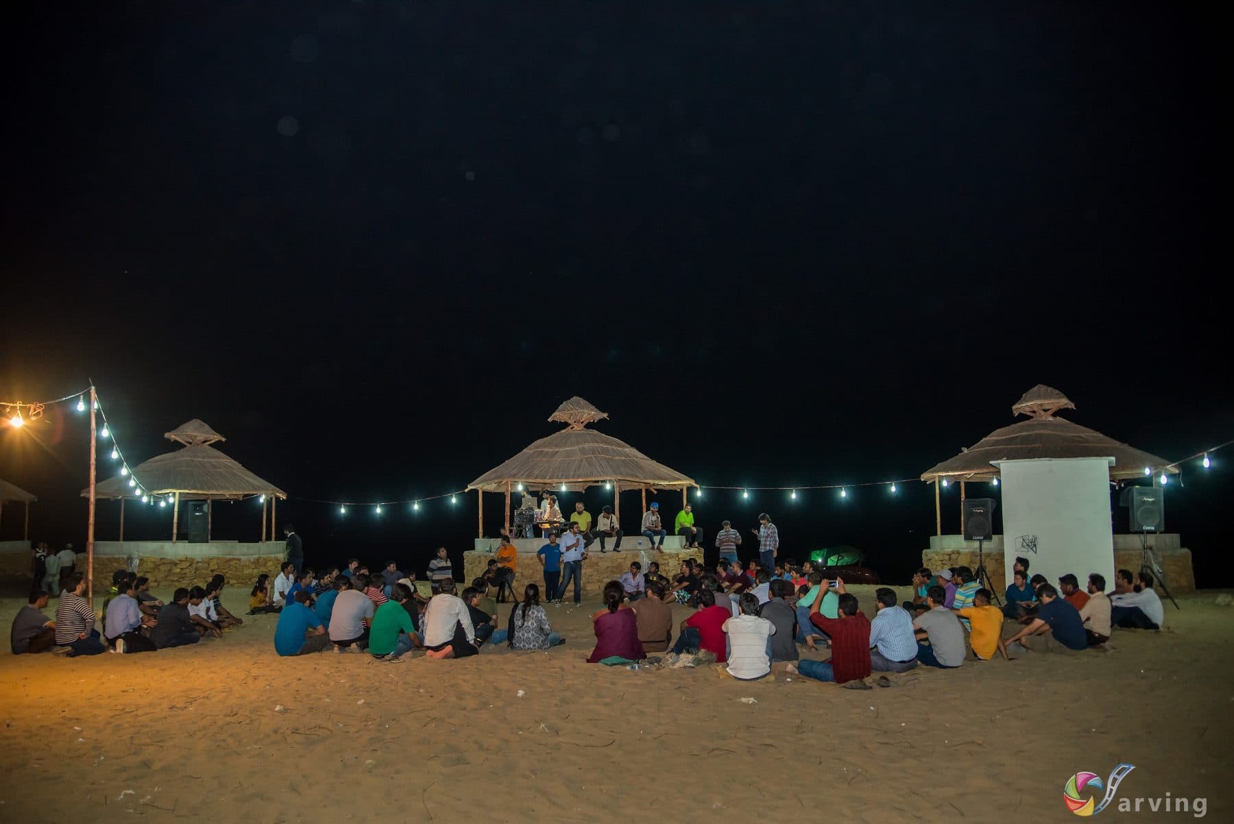 A retreat group seated in a wide circle outdoors after dark, strung lights overhead