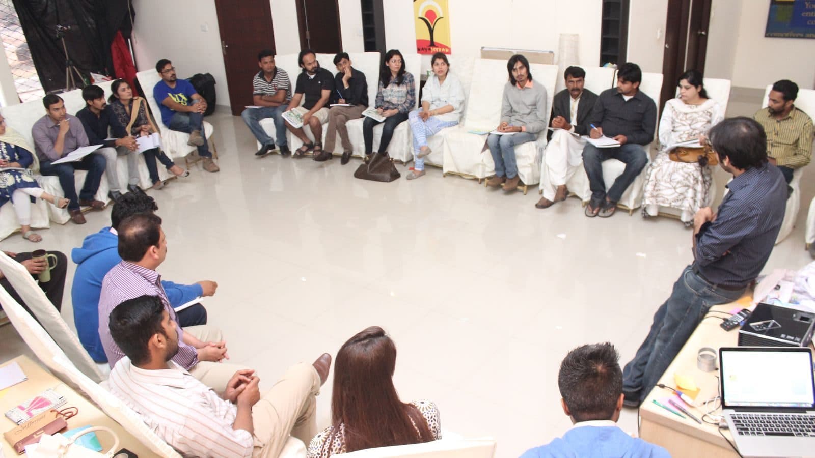 A wide circle of chairs in a bright room, everybody facing inward with notes on their laps