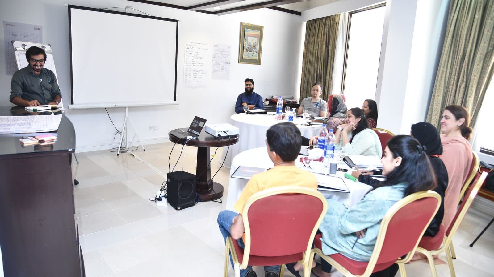 A training room with a projector screen at the front and participants working at tables along the walls