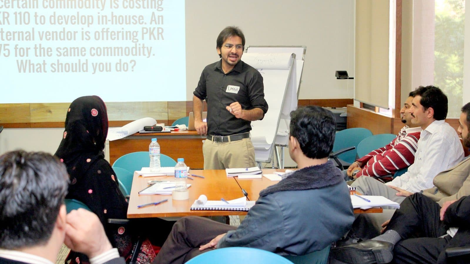 A facilitator at a flipchart with a costing problem up on the screen behind him and a table of participants working it