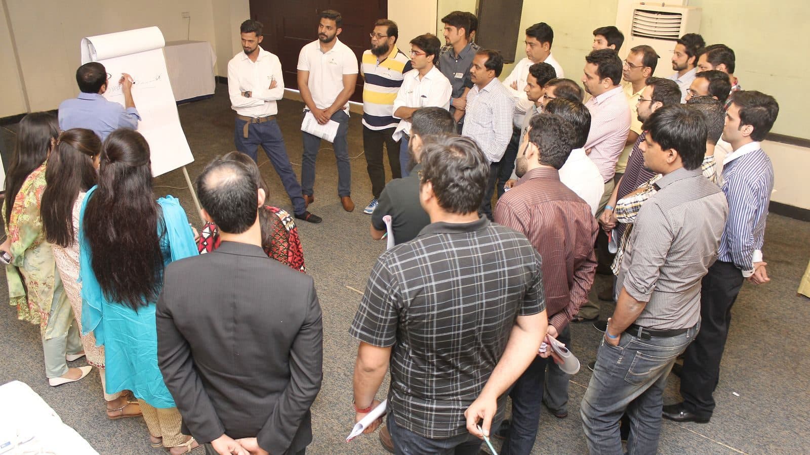 A large group standing in a loose ring around flipcharts propped on the floor
