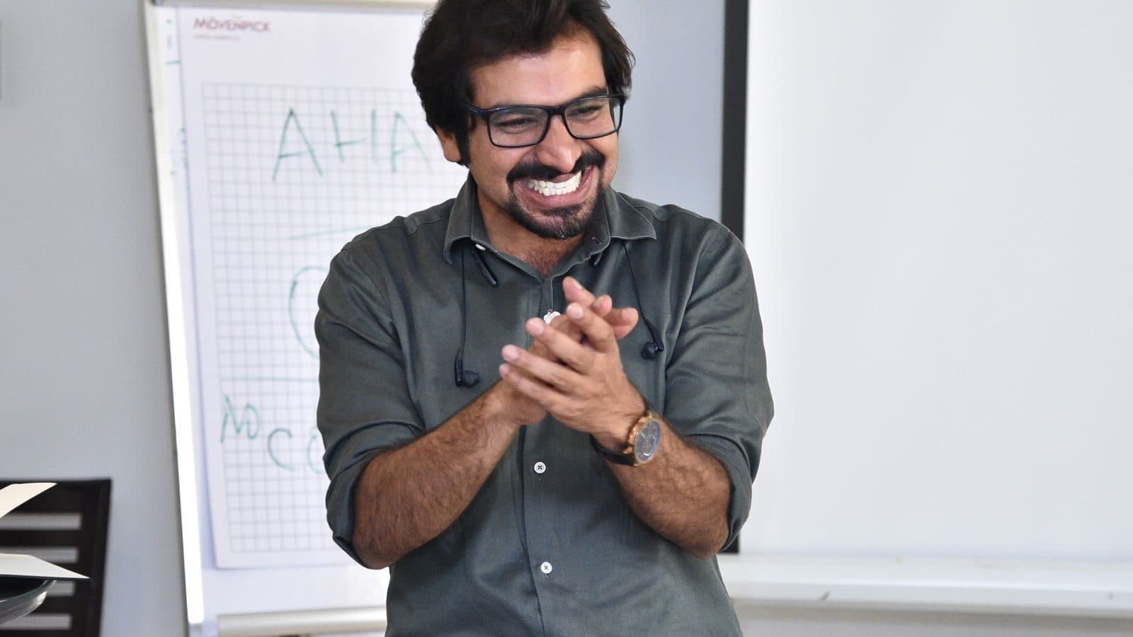 A trainer laughing with his hands together in front of a whiteboard